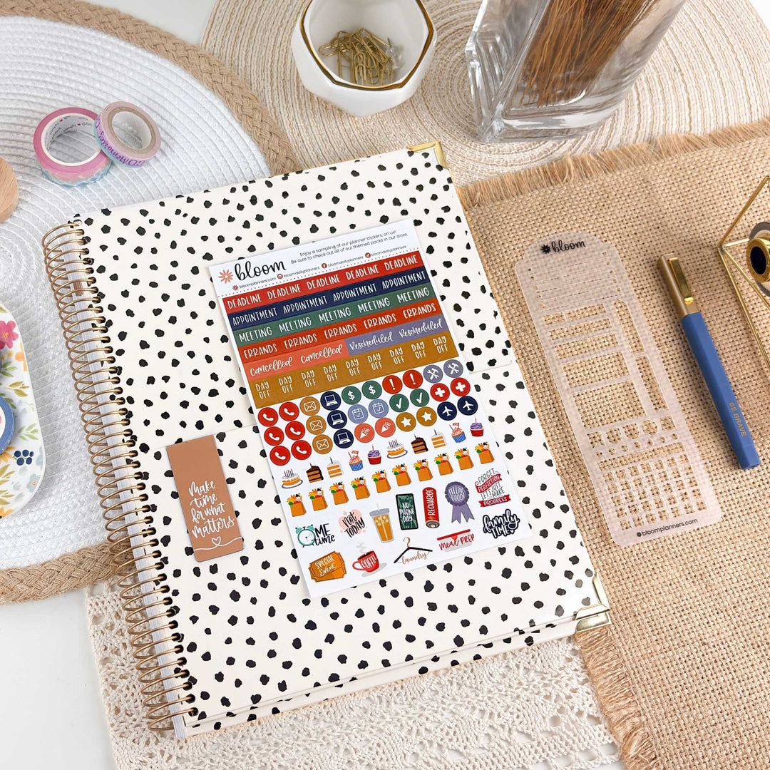 A spiral-bound planner or journal with a white cover covered in black polka dots, shown open and filled with colorful planner stickers. The item is photographed in a styled desk setup with burlap mats, washi tape, a pen, and other stationery accessories.