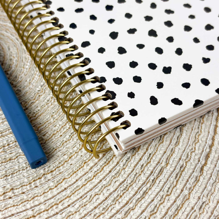 A spiral-bound notebook with a white cover and black irregular polka dots, shown with a gold-tone spiral binding on a textured beige woven surface. A blue pen is partially visible at the left edge.