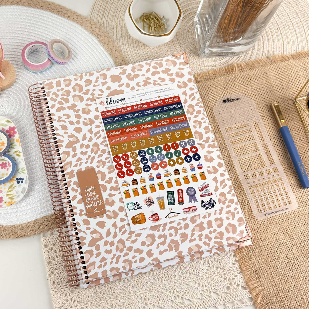 A spiral-bound planner or notebook with a beige and brown leopard print cover, decorated with colorful planner stickers. It is styled on a burlap mat with other desk accessories including washi tape, a glass of water, and a pen.