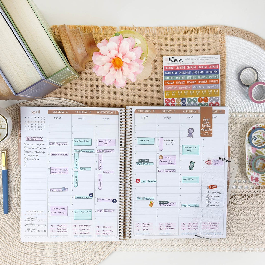 An open spiral planner shown from above on a woven mat, surrounded by decorative props including books, a pink flower, washi tape, and sticker sheets. The planner pages are filled with weekly and daily planning stickers and notes.