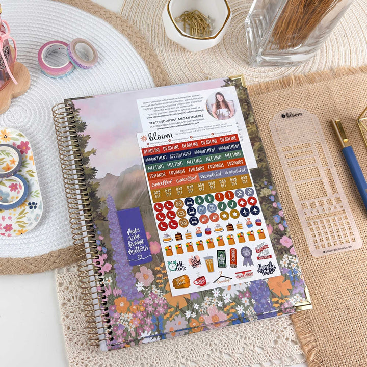 A spiral-bound planner or notebook with a colorful floral cover and a large sticker sheet attached, styled on a woven mat with desk accessories including washi tape, a pen, a clear grid sheet, and a toothpick holder.
