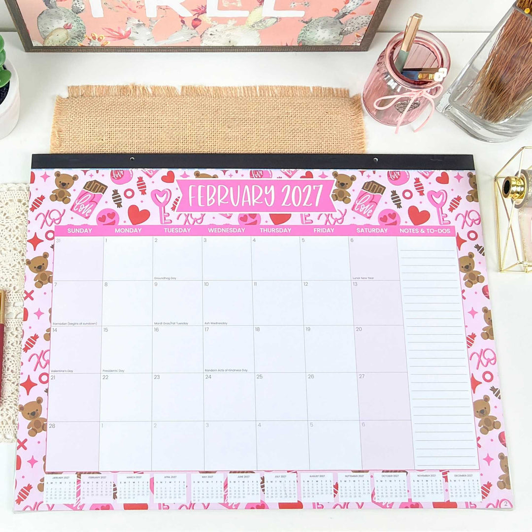 February 2027 calendar with teddy bear and heart designs on a desk.