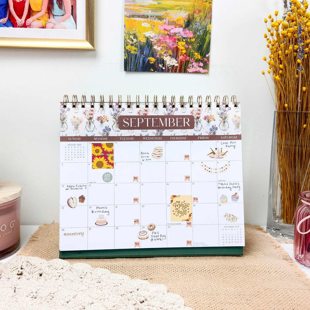 Hand writing in the September 2027 bloom Standing Desk Calendar with washi tape and gold accents nearby.