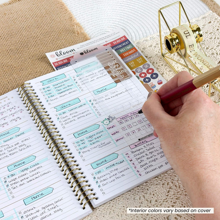 An open spiral planner with colorful weekly layout pages and stickers, being filled out by a hand holding a pen. A gold decorative object and a lace surface are visible in the background.