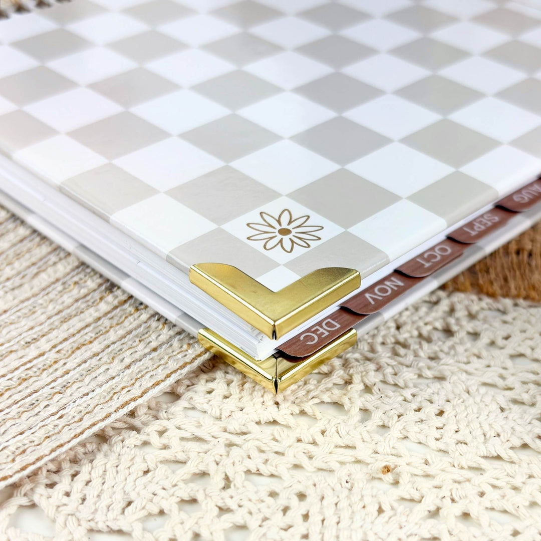 A close-up of a planner or notebook with a beige and white checkerboard cover, gold corner protectors, and visible monthly tabs labeled DEC, NOV, OCT, and NOV on a textured cream lace surface.
