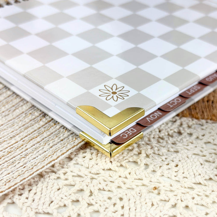 A close-up of a planner or notebook with a beige and white checkerboard cover, gold corner protectors, and visible monthly tabs labeled DEC, NOV, OCT, and NOV on a textured cream lace surface.