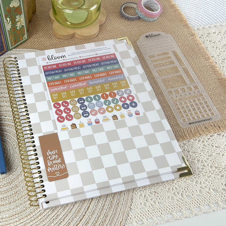 A spiral-bound planner sticker sheet set from the brand bloom, featuring a beige and white checkered cover with various colorful planner stickers and a brown accent sticker. The planner is styled on a textured beige mat alongside a clear plastic ruler and washi tape.