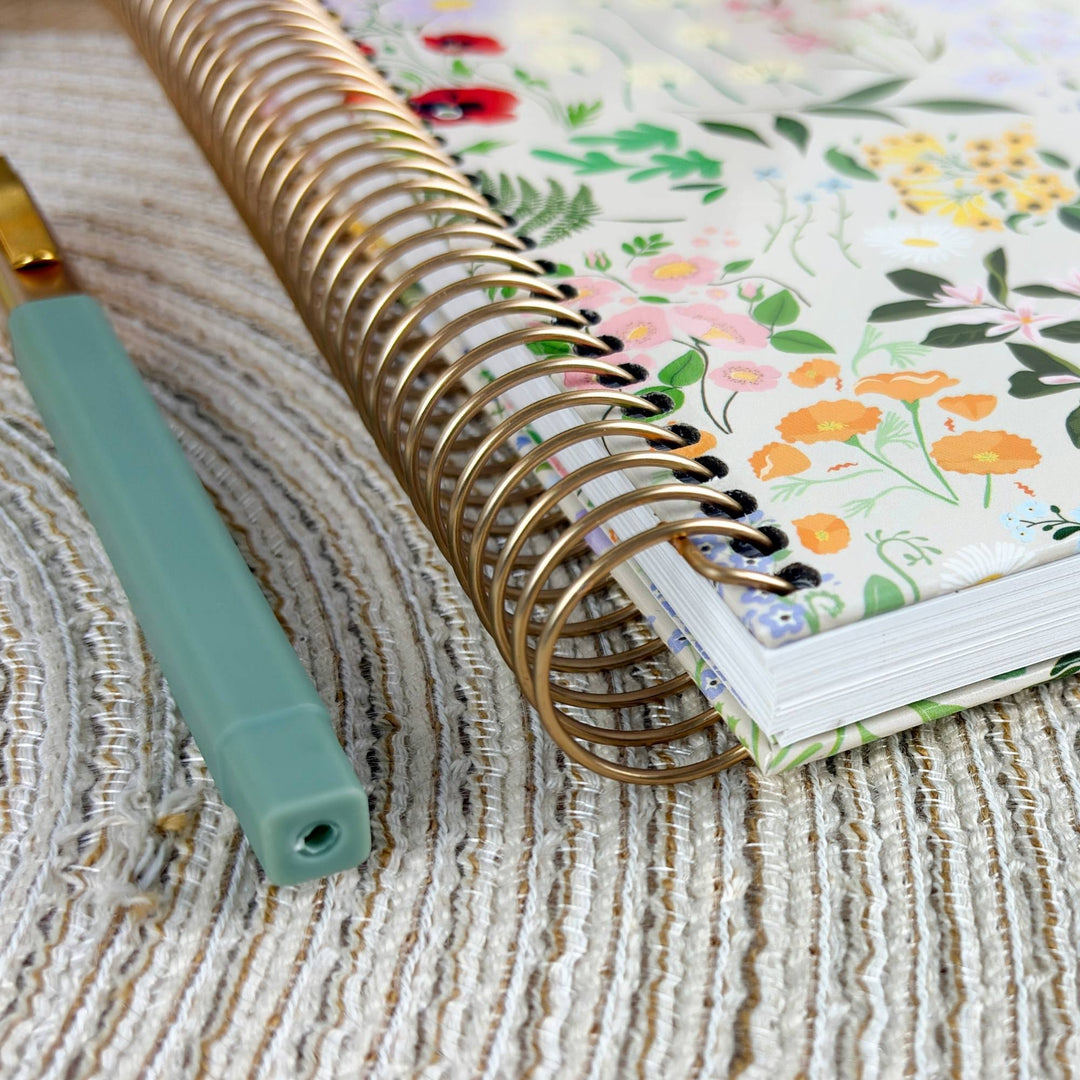 A close-up of a floral-patterned spiral notebook with a gold wire binding, placed on a textured beige woven surface next to a mint green pen.