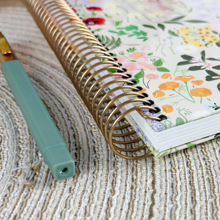 A close-up of a floral-patterned spiral notebook with a gold wire binding, placed on a textured beige woven surface next to a mint green pen.