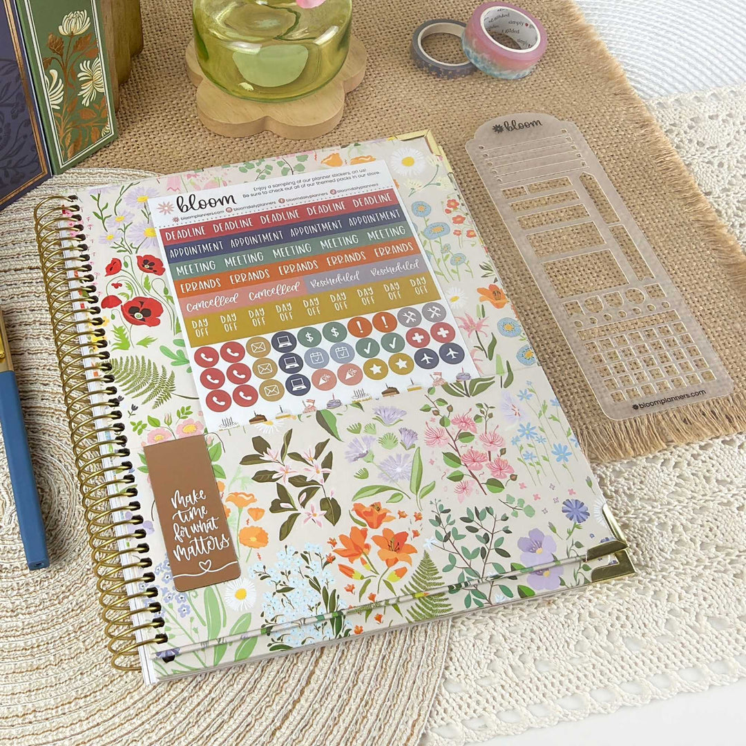 A spiral-bound planner decorated with a colorful floral sticker layout, shown on a woven beige surface with coordinating stationery props including washi tape, a clear grid sticker sheet, and a green candle.