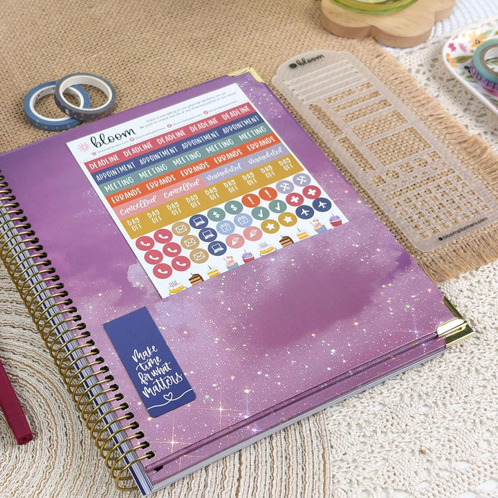A spiral-bound planner with a purple glittery cover and colorful sticker sheets, styled on a beige woven mat with washi tape and a clear ruler nearby.