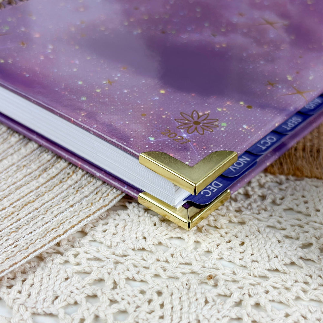 A close-up of a purple glittery planner or notebook cover with gold foil details, held open by a gold metal L-shaped corner bookmark on white pages, resting on a cream lace textile.