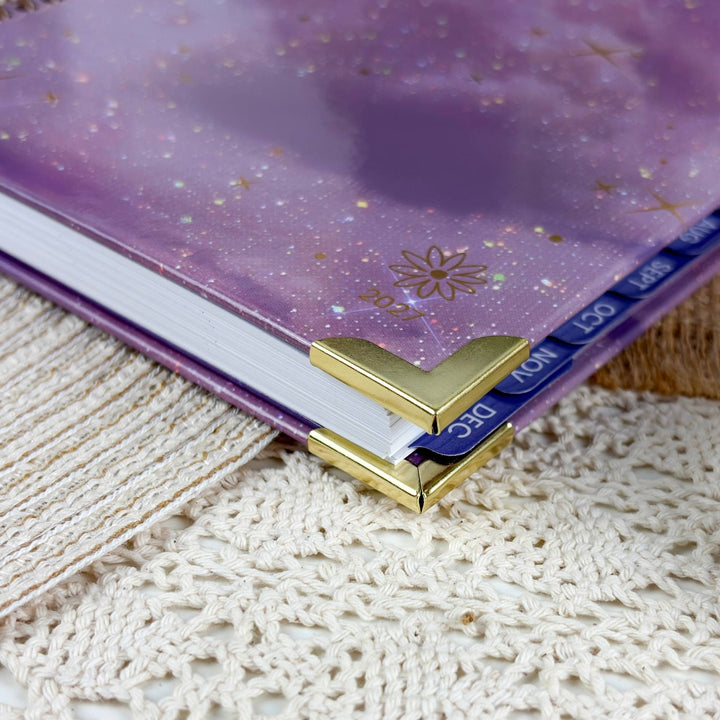 A close-up of a purple glittery planner or notebook cover with gold foil details, held open by a gold metal L-shaped corner bookmark on white pages, resting on a cream lace textile.