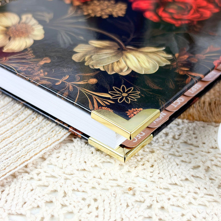 A close-up of a decorative book or journal with a glossy floral cover, featuring large cream and red flowers on a dark background, with gold corner protectors and a textured cream lace surface underneath.
