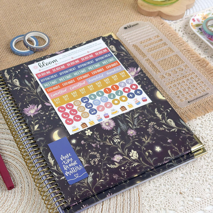 A spiral-bound planner or notebook with a dark floral cover lies on a woven mat, surrounded by stationery props including a clear plastic divider, washi tapes, and a small dish of colorful items. The cover has a black base with gold-toned floral linework, pink flowers, and small moon/star motifs.