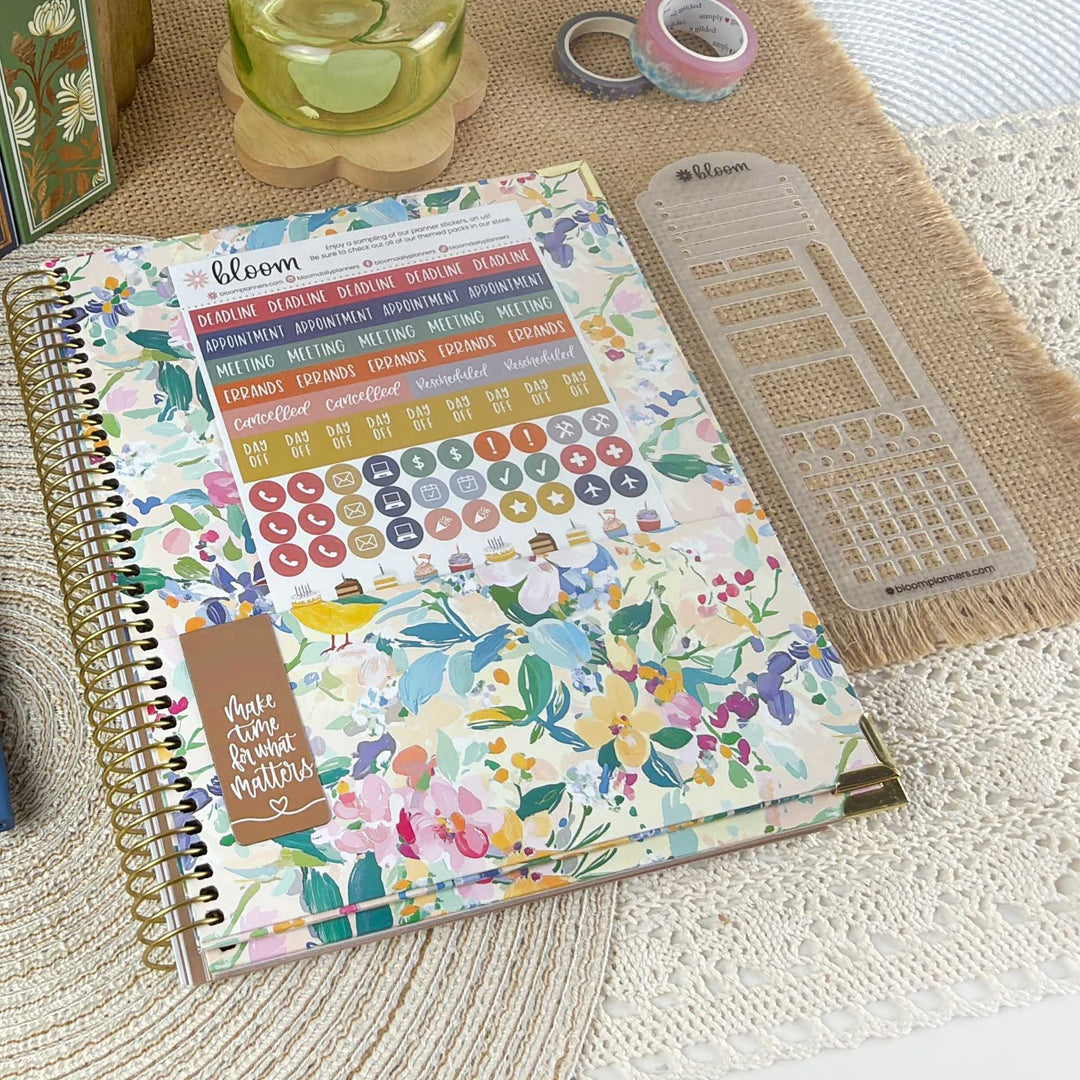 A floral planner or journal cover with a gold spiral binding, shown on a woven mat with planner stickers and a clear grid template nearby. The cover features a multicolor watercolor floral pattern with the brand name 'bloom' visible.