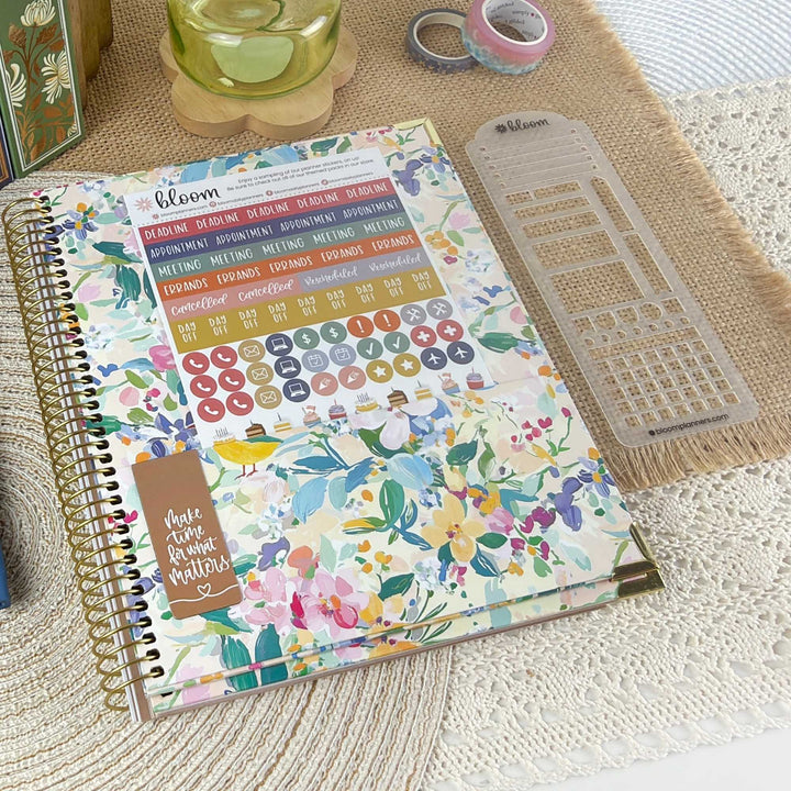 A floral planner or journal cover with a gold spiral binding, shown on a woven mat with planner stickers and a clear grid template nearby. The cover features a multicolor watercolor floral pattern with the brand name 'bloom' visible.