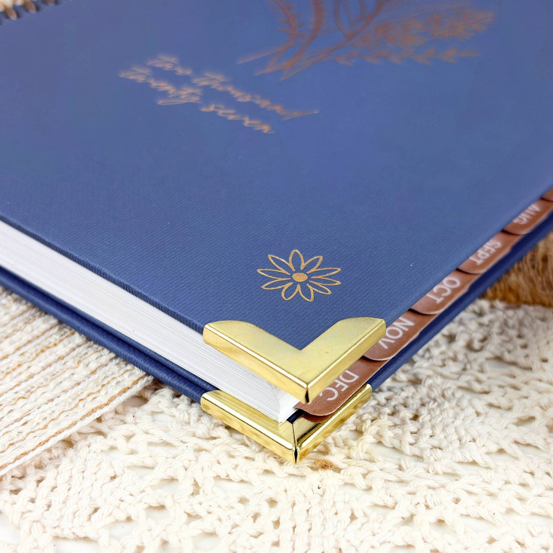 A close-up of a blue hardcover journal or planner with gold foil floral details, white pages, and gold corner protectors, resting on a cream textured fabric surface.