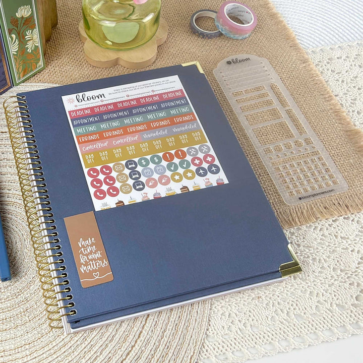 A navy blue spiral-bound planner or notebook with a decorative sticker sheet attached to the cover, styled on a woven beige mat with other stationery items nearby.