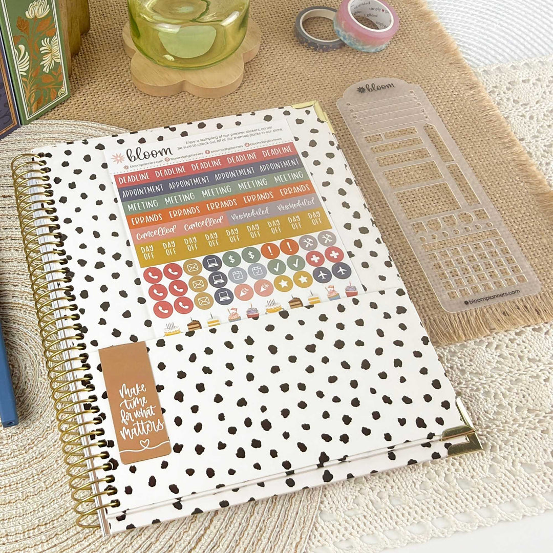 A spiral-bound planner or notebook with a white cover covered in black heart print, shown open with colorful sticker sheets and a clear plastic grid insert. The item is styled on a beige textured surface with decorative props.