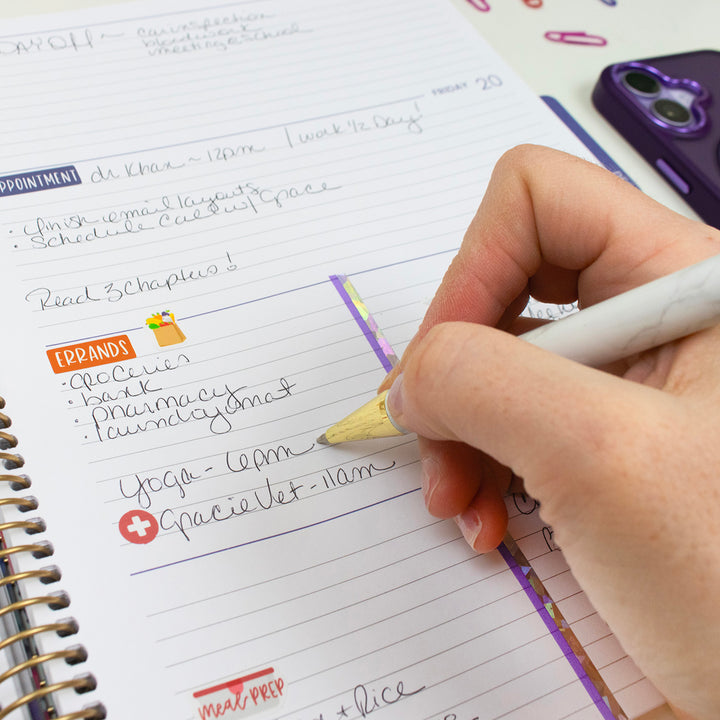 Hand writing in a planner with a pencil, surrounded by stationery items.