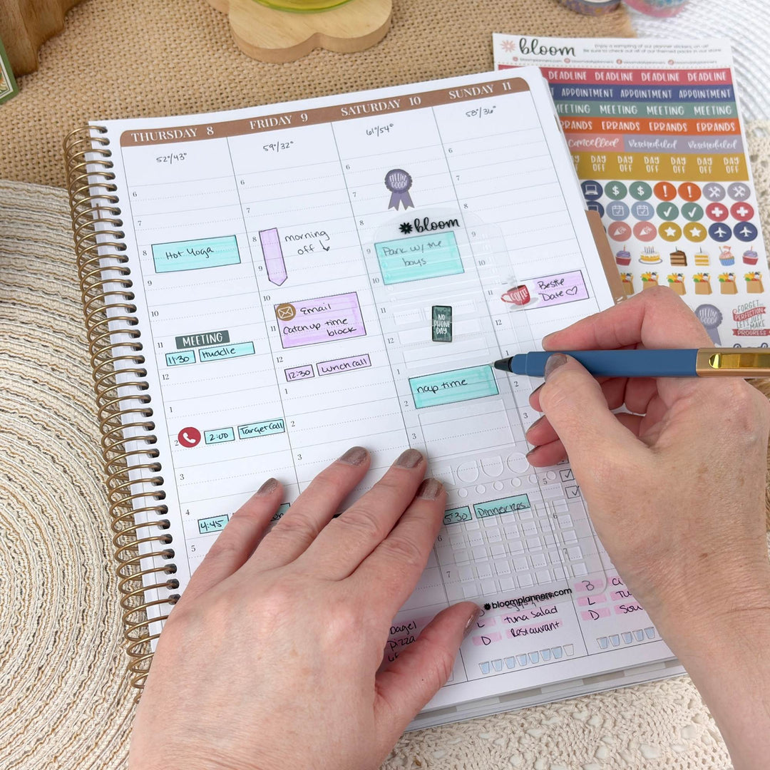 A weekly planner spread open on a woven mat, with a hand holding a blue pen writing in the pages. The planner has a white page with brown weekday headers and colorful planner stickers, and a sheet of matching stickers is visible at the top right.