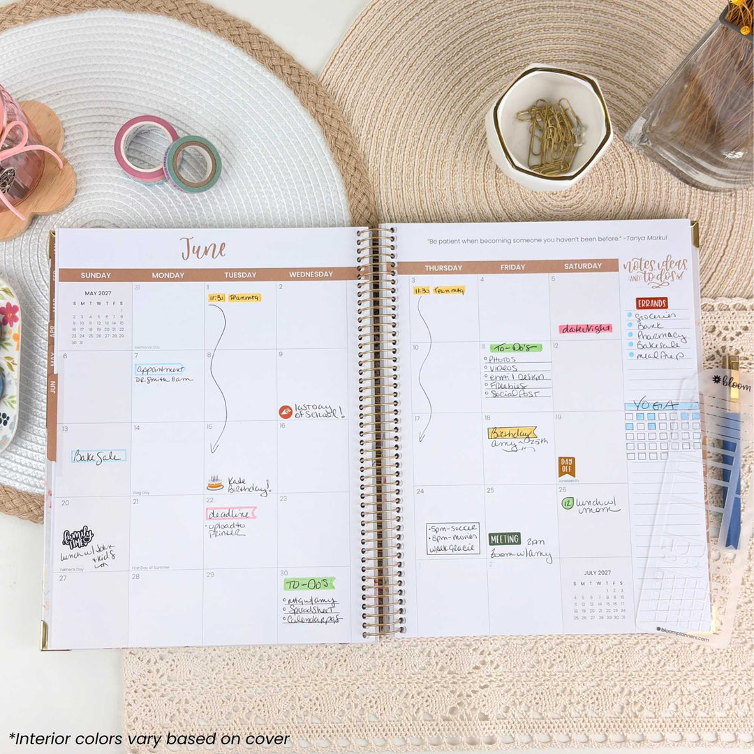 An open spiral-bound planner showing the June 2023 weekly calendar spread, styled on a woven placemat with pastel washi tape, a small dish of paper clips, and a glass bottle nearby. The planner pages are white with tan and gold accents and handwritten notes in multiple colors.