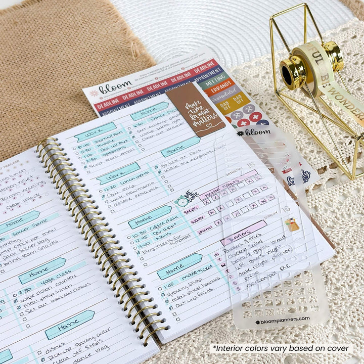 An open spiral planner showing decorated weekly pages, with a roll of planner stickers on a gold spool and a branded sticker sheet nearby. The scene is styled on a lace doily over burlap fabric.