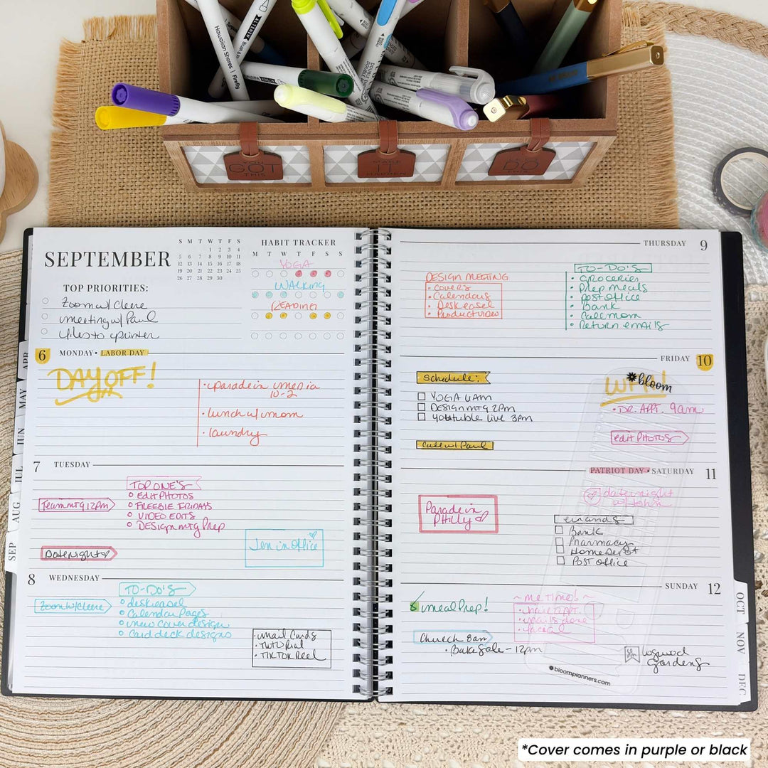 An open spiral-bound planner showing a September weekly layout with handwritten notes in multiple colors. A wooden organizer filled with pens and markers sits behind it on a woven surface.
