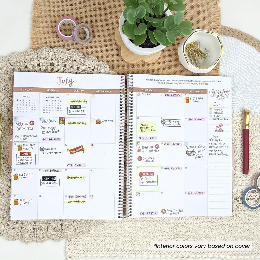 An open spiral-bound planner showing a July calendar spread, styled on a beige woven mat with a small potted succulent, decorative rings, a gold dish, and a pen around it.