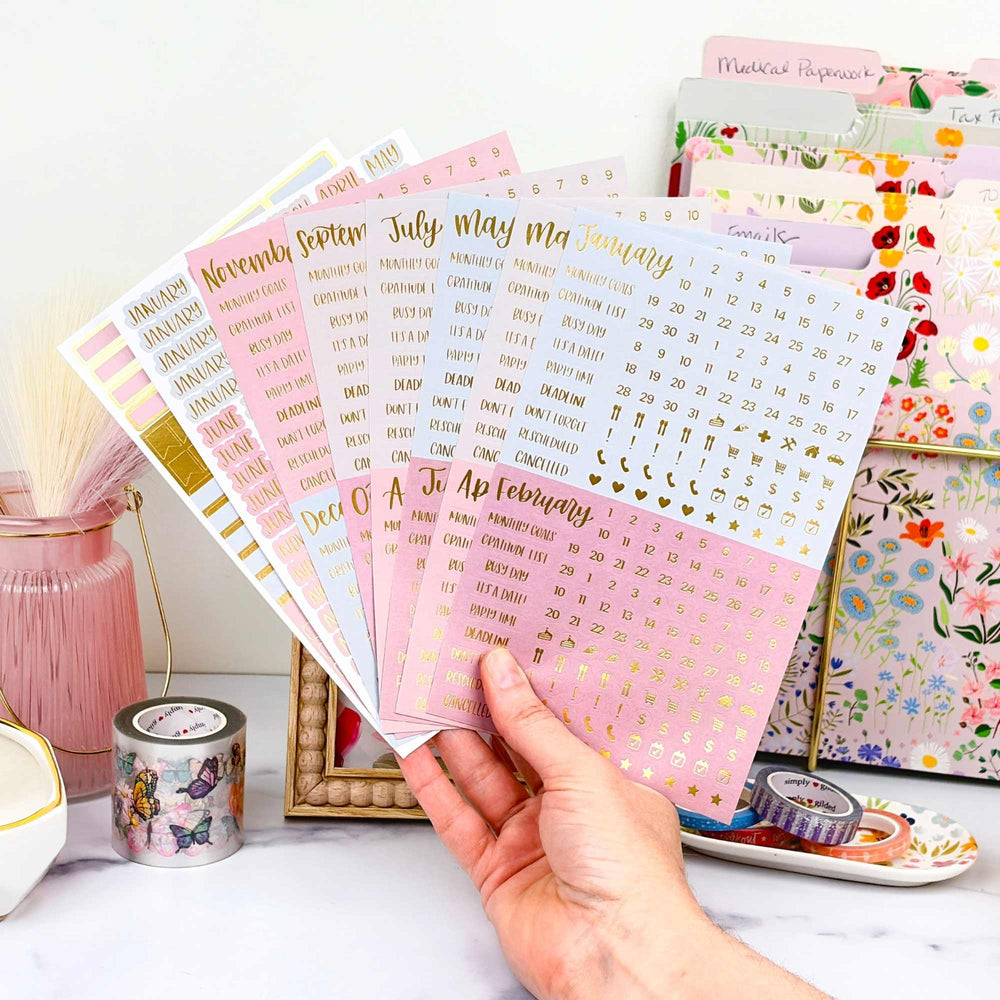 Hand holding a stack of decorative calendar essential planner stickers in matte jewel tone colors. - hover