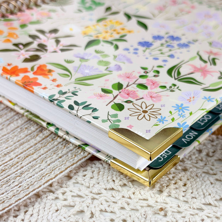 A close-up of a floral-patterned notebook or planner cover with gold metal corner protectors and visible monthly tabs, resting on a textured cream woven surface.