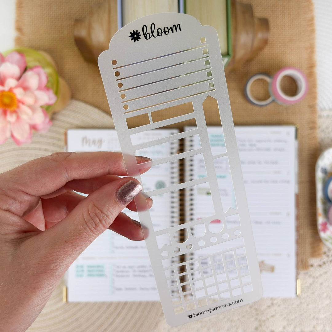 A hand holds a white plastic planner stencil with the brand name 'bloom' printed at the top. The stencil features various grid and slot cutouts.
