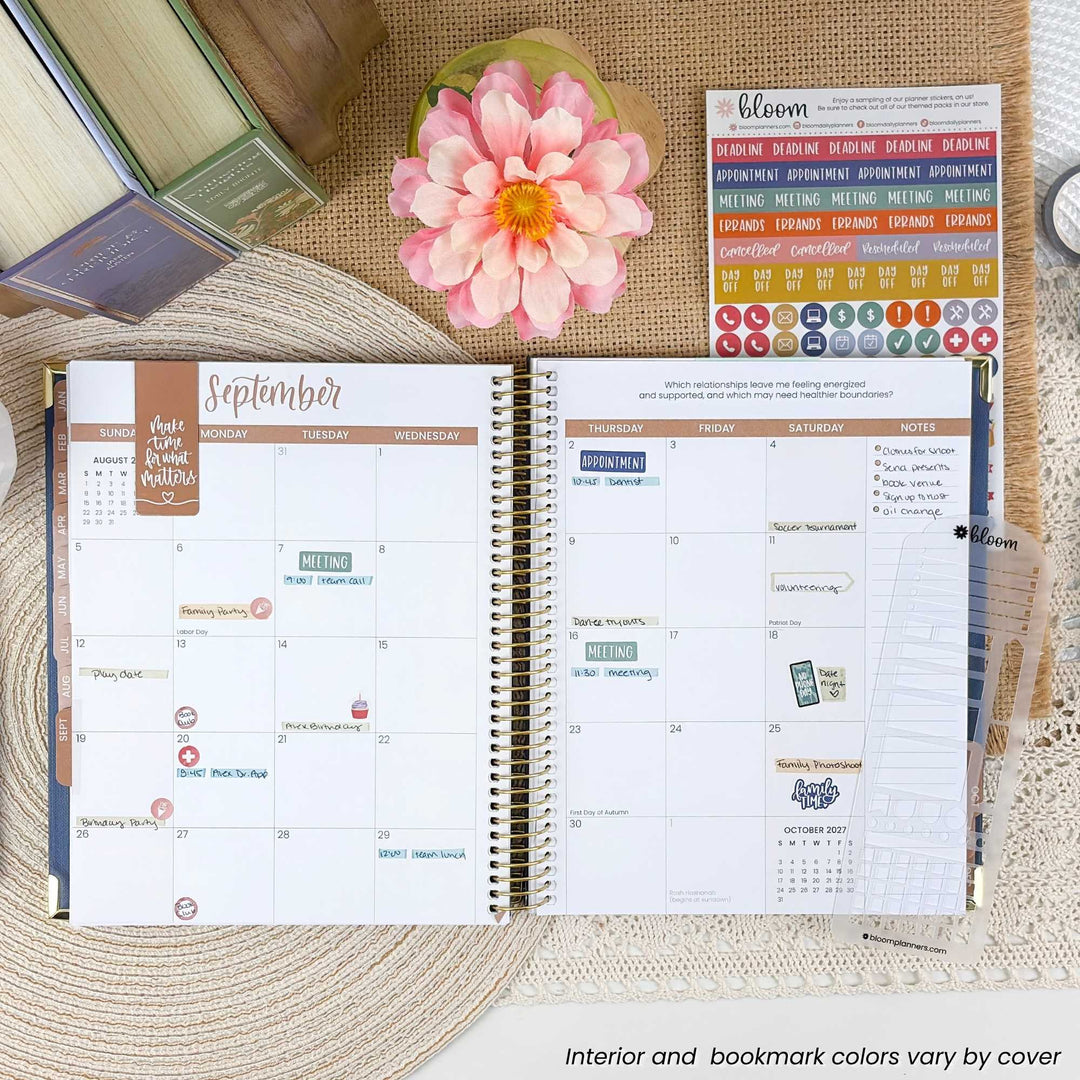 An open spiral-bound planner showing a September calendar spread with stickers and bookmarks, styled on a woven mat with a pink flower, books, and a small card in the background.