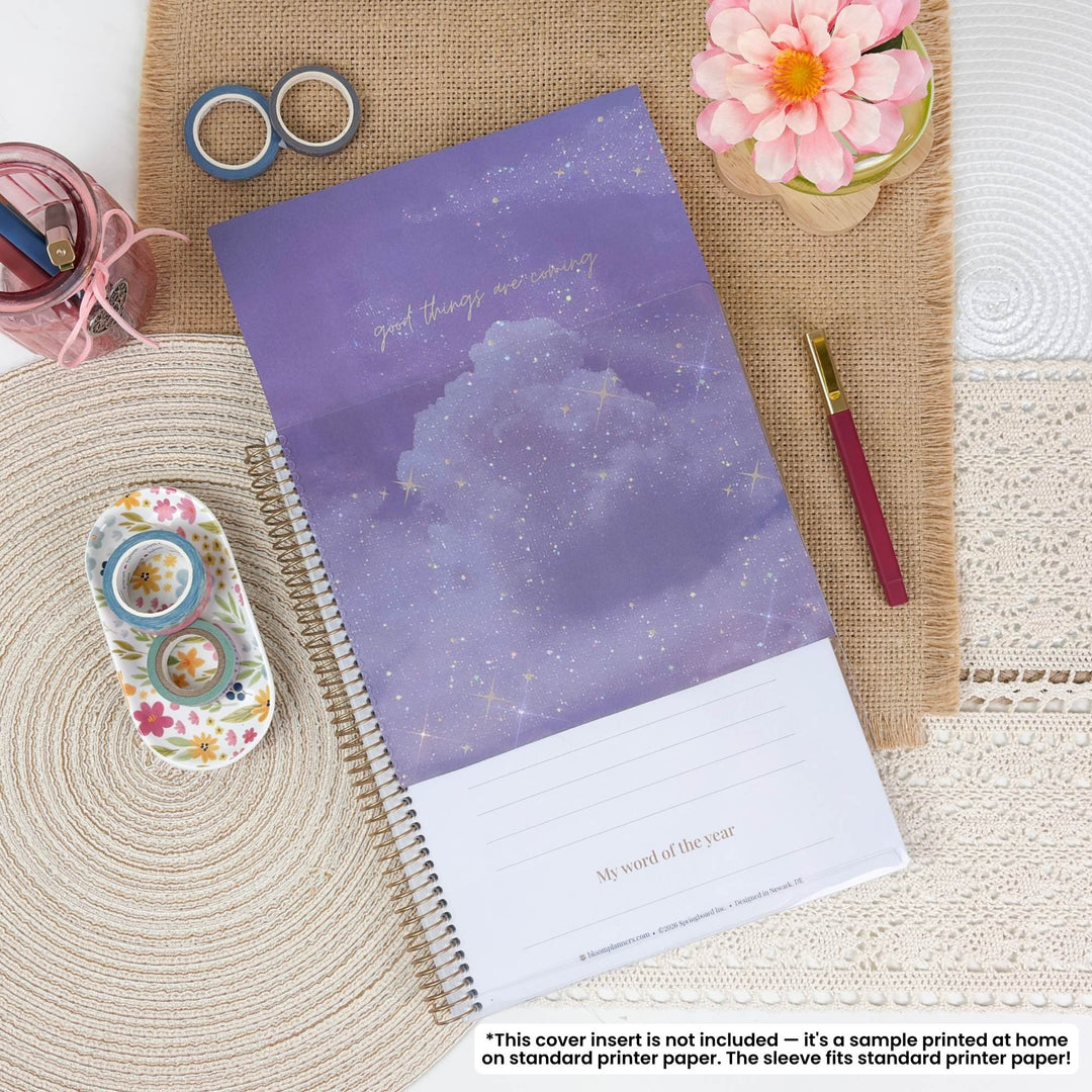 A spiral-bound planner or notebook with a purple cloud and starry sky cover design, styled in a flat lay on burlap and lace with washi tape, a pen, flowers, and stationery accessories. A small disclaimer note appears at the bottom.