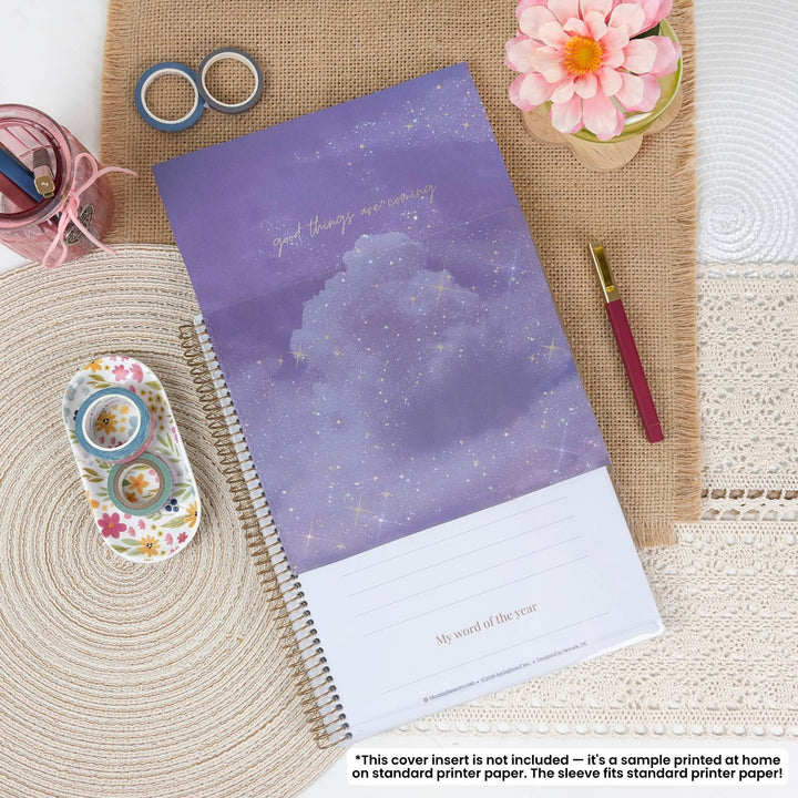 A spiral-bound planner or notebook with a purple cloud and starry sky cover design, styled in a flat lay on burlap and lace with washi tape, a pen, flowers, and stationery accessories. A small disclaimer note appears at the bottom.