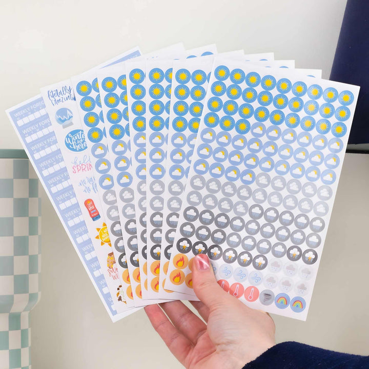 Hand holding a set of decorative weather planner stickers with various weather related icons on a neutral background