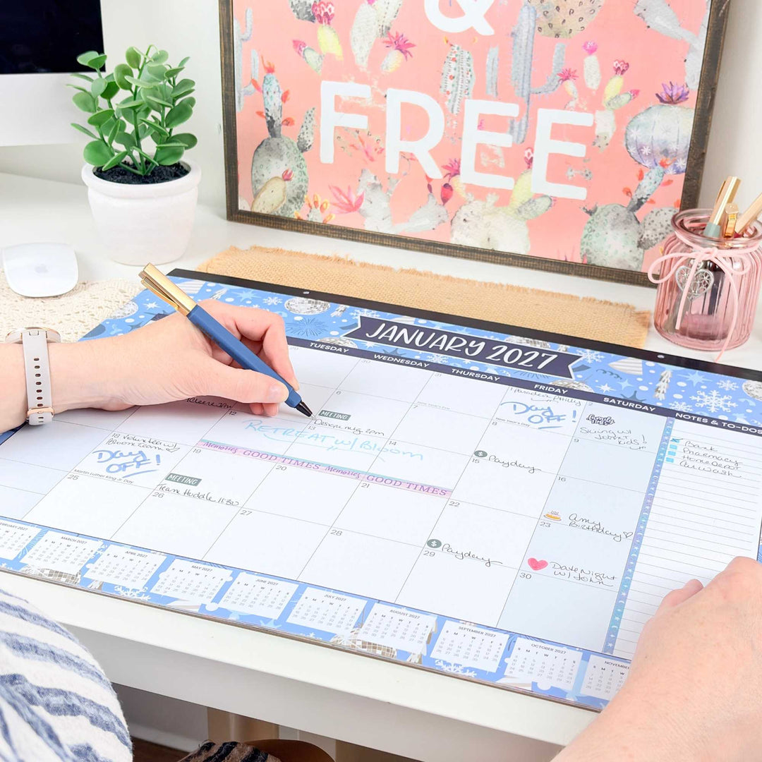 Person using a large desk calendar with a decorative plant and framed picture in the background.