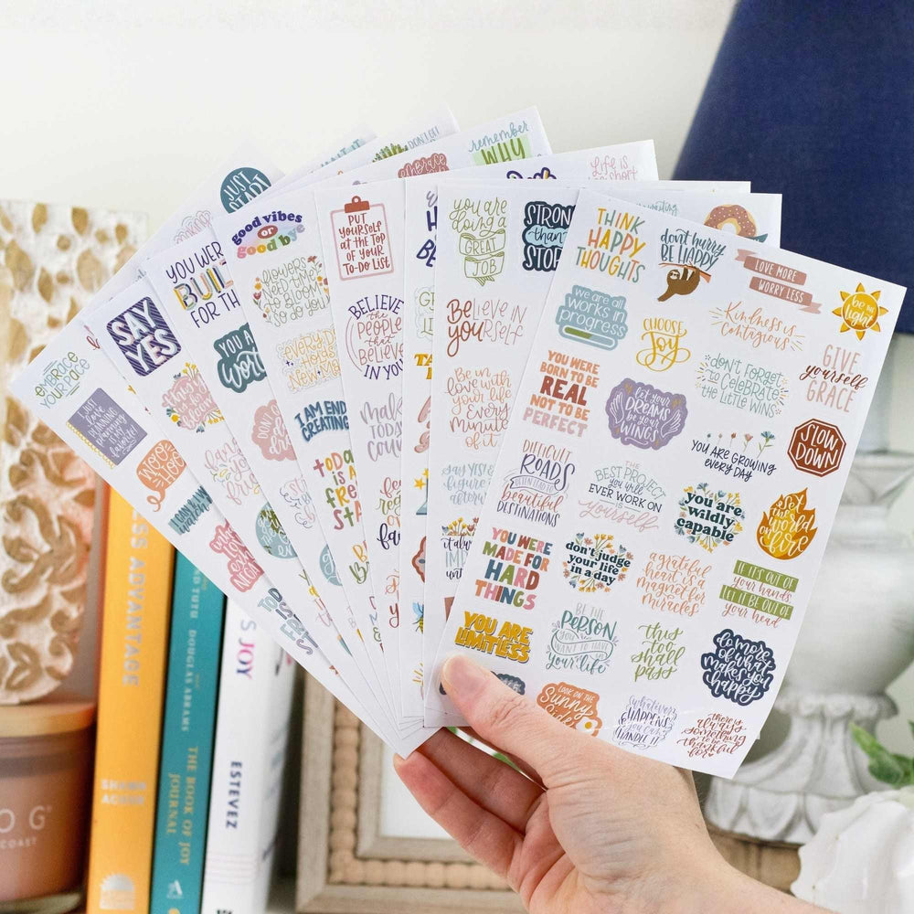 Hand holding a stack of motivational sticker sheets with colorful text and graphics.