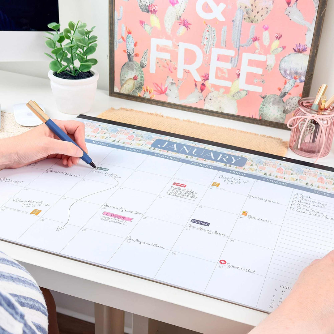 Person using a pen to mark on a large calendar on a desk with a plant and decorative items in the background.