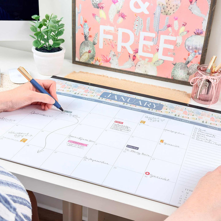 Person using a pen to mark on a large calendar on a desk with a plant and decorative items in the background.