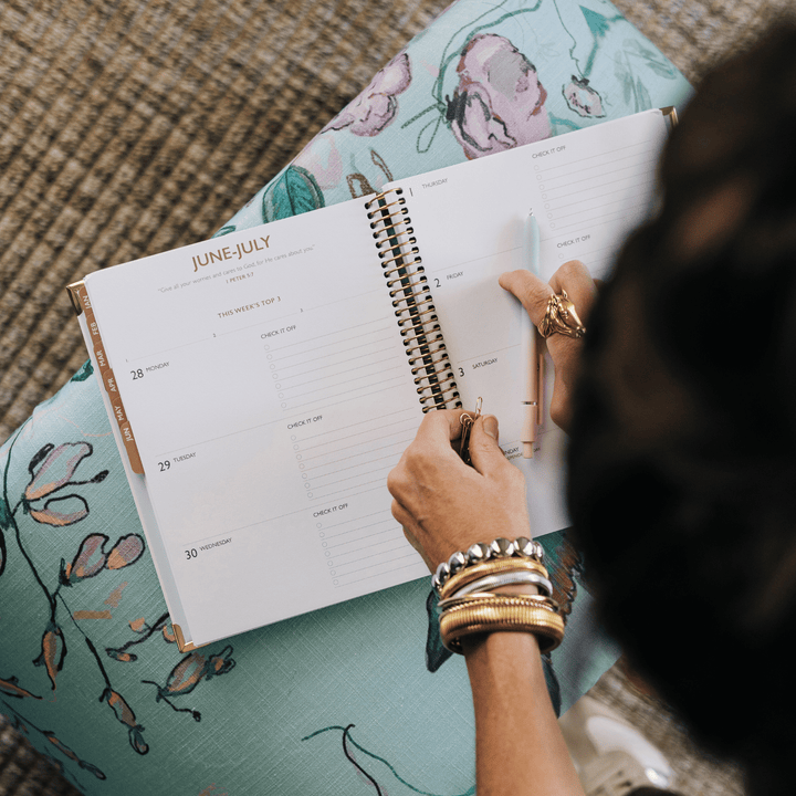 An open spiral-bound planner or calendar lies on a person's lap, showing a June-July weekly layout. A hand holds a pen over the pages, with stacked bracelets visible on the wrist.