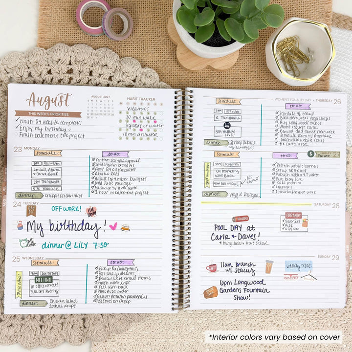 An open spiral-bound planner shown from above on a beige textured surface with a crocheted lace cloth underneath. The planner pages are filled with handwritten notes, weekly layouts, and colorful decorative accents.