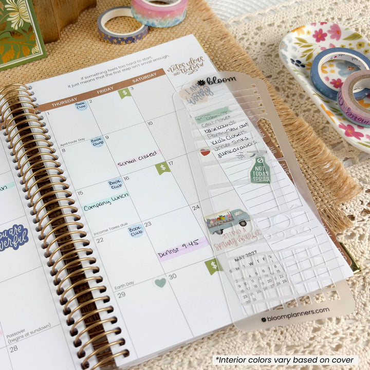A spiral-bound planner open to a weekly calendar page, decorated with a clear plastic cover and small decorative stickers. The scene is styled on a textured beige surface with washi tape rolls and a floral dish in the background.