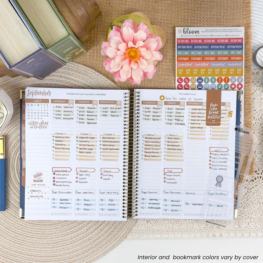 An open spiral-bound planner spread showing a detailed monthly calendar layout with columns for tasks, meetings, and notes. The planner is styled on a woven placemat with a pink flower, books, and sticker sheets around it.