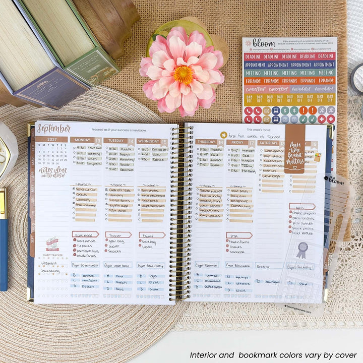 An open spiral-bound planner spread showing a detailed monthly calendar layout with columns for tasks, meetings, and notes. The planner is styled on a woven placemat with a pink flower, books, and sticker sheets around it.