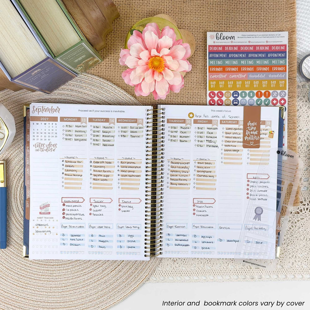 An open spiral-bound planner spread showing a detailed monthly calendar layout with columns for tasks, meetings, and notes. The planner is styled on a woven placemat with a pink flower, books, and sticker sheets around it.