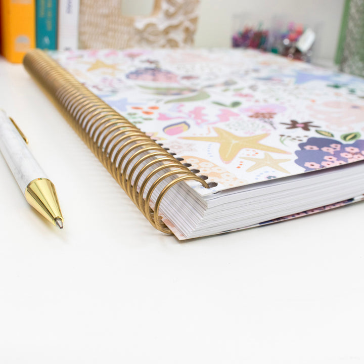 Planner with floral design on cover and gold pen on white surface