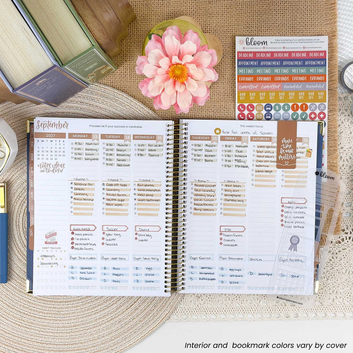 An open spiral-bound planner spread showing a detailed monthly calendar layout with columns for tasks, meetings, and notes. The planner is styled on a woven placemat with a pink flower, books, and sticker sheets around it.