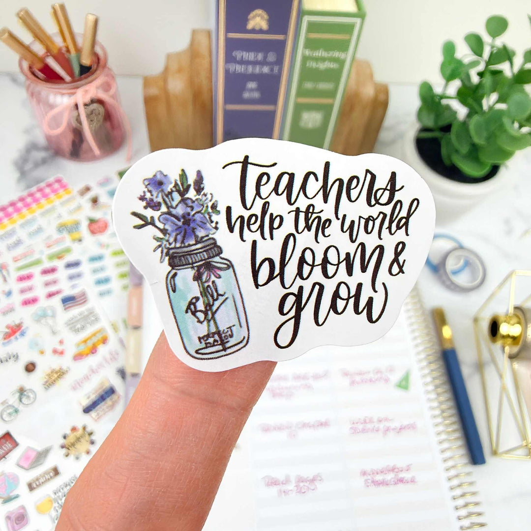 Sticker with 'Teachers help the world bloom & grow' text held over a desk with stationery items.